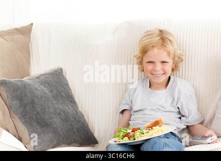Child boy holding salad plate and TV remote while sitting on sofa at home, copy space Stock Photo