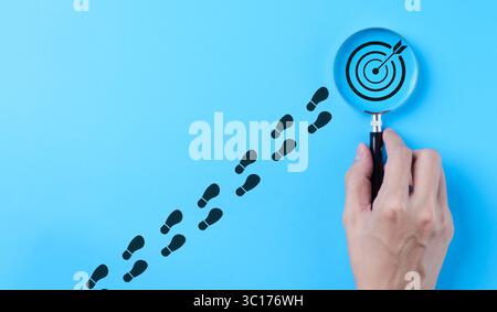 Hand holding magnifying glass focusing on target goal icon on blue background. Business achievement goal and objective target concept. Growth Stock Photo