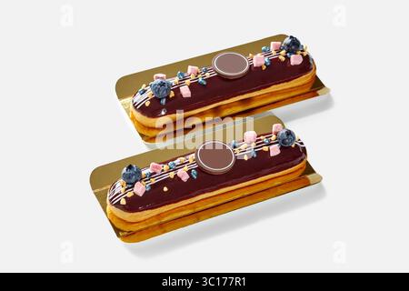 Two chocolate eclairs decorated with blueberries and sprinkles resting on golden trays Stock Photo