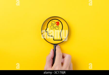 Hand holding magnifying glass focusing on human brain jigsaw puzzle on yellow background. human psychology or mental health or illness, idea, brainsto Stock Photo