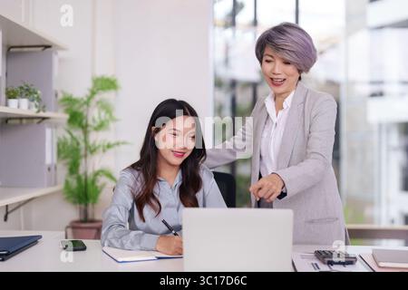 Asian businesswomen mentor young professional learning skills. Team collaboration in a modern office. Experienced leader guiding career growth. Stock Photo