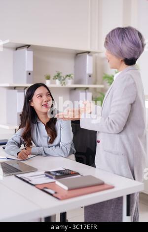 Two Asian women engage in a productive business discussion in a contemporary office setting. Mentorship and professional growth are evident. The scene Stock Photo