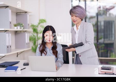 Two Asian businesswomen collaborating digitally in contemporary office. Experienced mentor guides young professional on computer tasks. Stock Photo