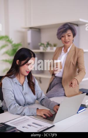 Two Asian professional women engage in productive office collaboration. An experienced mentor assists a young colleague reviewing data on a laptop in Stock Photo