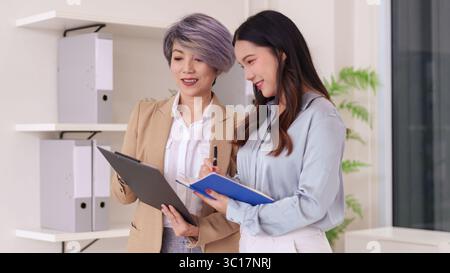 Two diverse Asian females engaged in productive office discussion. Experienced mentor guiding young professional in a modern workplace setting. Stock Photo