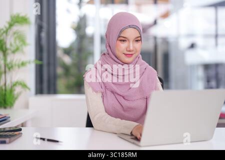 Young Asian Muslim female expert typing on a laptop at her bright desk focused on professional tasks and online career development. Stock Photo