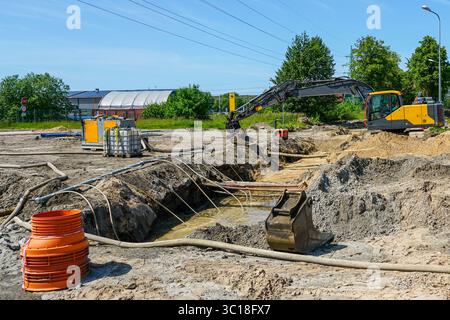 Foreground shows drainage system with pump, hoses, and pipes managing trench water, excavator works behind Stock Photo