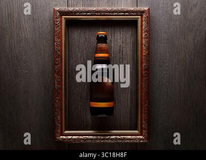 A bottle of dark beer is suspended within an ornate, gilded picture frame. The frame is mounted on a dark, textured wooden wall. The bottle has a brow Stock Photo