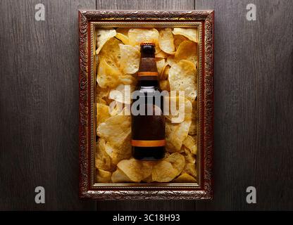 A dark brown wooden frame contains a bottle of dark beer and a pile of golden potato chips. The frame has a gold-colored border. The chips are arrange Stock Photo