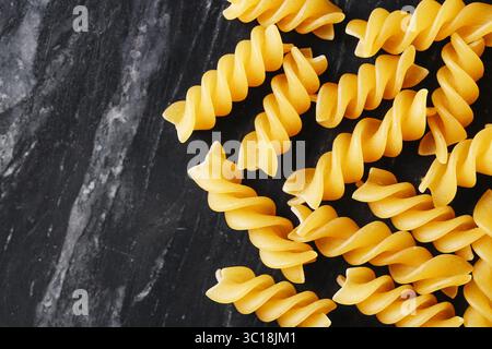 Raw pasta fusilli on dark stone. Yellow noodles spread on marble surface. Italian food concept. Dry spiral shapes close up. Culinary ingredient. Stock Photo