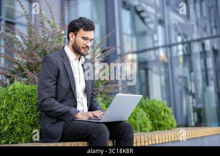 A businessman in glasses works on his laptop outdoors near a modern building and greenery. Stock Photo