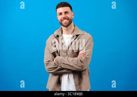 Bearded man with arms crossed wearing beige jacket against blue background Stock Photo