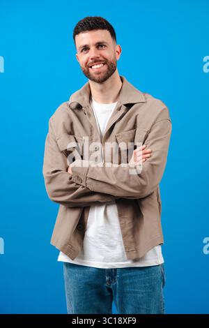 Bearded man with arms crossed wearing beige jacket against blue background Stock Photo