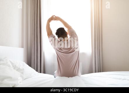 Back view of man wearing pajamas sitting on bed, stretching Stock Photo