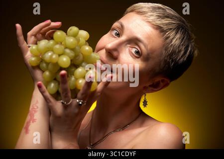 A young woman with short blonde hair holds a large bunch of green grapes in front of her face, looking slightly surprised and playful. She is wearing Stock Photo