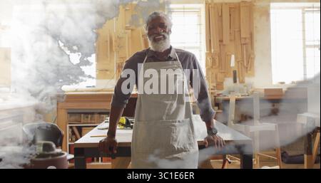 Standing African American carpenter wearing gray apron sanding wooden plank in carpentry workshop Stock Photo