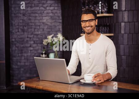 Man working on silver laptop and holding coffee cup at wooden table in industrial cafe space Stock Photo
