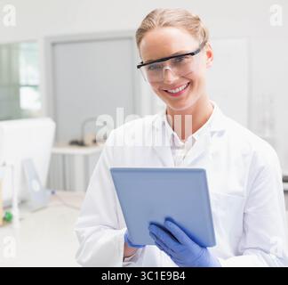 Female lab technician wearing white coat, safety goggles, gloves, holding tablet among lab benches Stock Photo