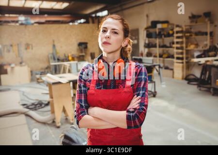Female woodworker wearing red apron and earmuffs, standing amid workbenches and lumber in workshop Stock Photo