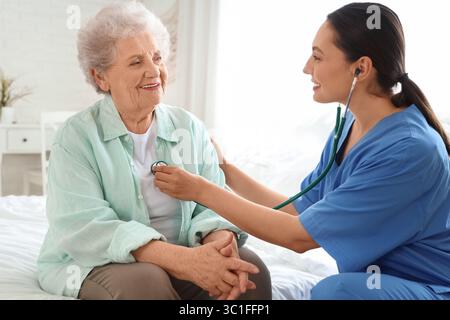 Physical therapist listening to senior woman with stethoscope in bedroom Stock Photo