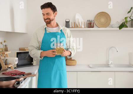 Handsome bearded man with jar of pasta and frying pan on stove in kitchen Stock Photo