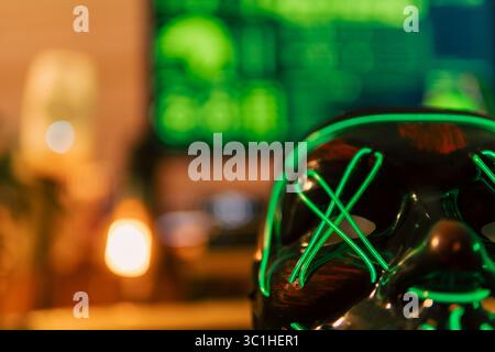 Neon-green hacker mask rests center stage on a cyber desk, while three widescreen monitors overflow with rapidly scrolling code. Stock Photo