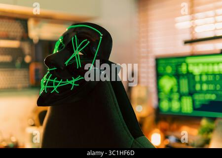 Neon-green hacker mask rests center stage on a cyber desk, while three widescreen monitors overflow with rapidly scrolling code. Stock Photo