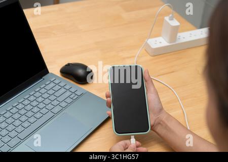 A person holding a smartphone and connecting the charging cable next to a laptop and mouse on a wooden office desk, showing modern technology and work Stock Photo