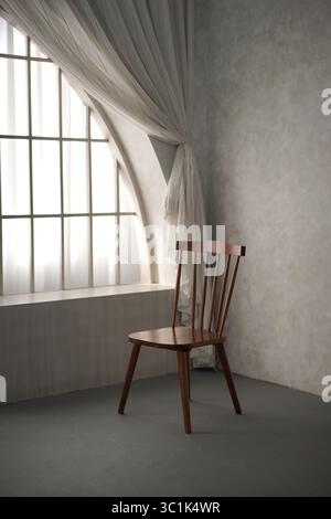 A single dark brown wooden chair in an empty room by a window with a white sheer curtain. Minimalist and elegant interior design with natural light. Stock Photo