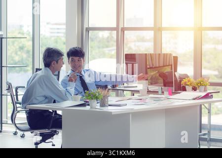 Two Asian businessmen in a bright office discussing work displayed on a desktop computer. They are collaborating and pointing at the screen. Stock Photo