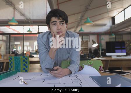 Native American man in gray shirt leaning over desk examining floor plan using smartphone at office Stock Photo