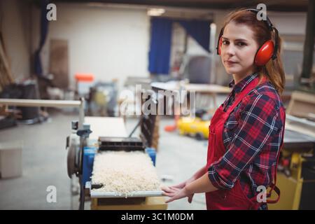 Female carpenter with red apron and earmuffs using planer in workshop making shavings, copy space Stock Photo
