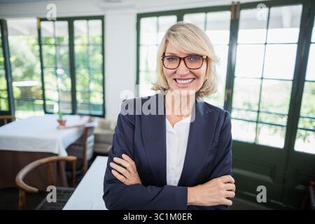 Mature woman wearing navy blazer and brown eyeglasses standing in dining area showing wooden chairs Stock Photo