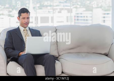 Middle-aged man typing on silver laptop sitting on sofa in office lounge wearing suit, copy space Stock Photo