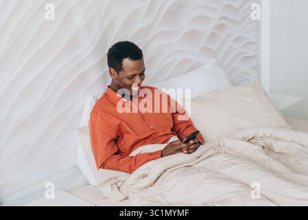 Happy african american man lying comfortably in a cozy bed, wrapped in a warm blanket while using a smartphone in a modern bedroom, enjoying a relaxing morning at home Stock Photo