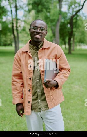 Portrait of Young Adult Black Man Smiling and Holding Laptop in Park Stock Photo
