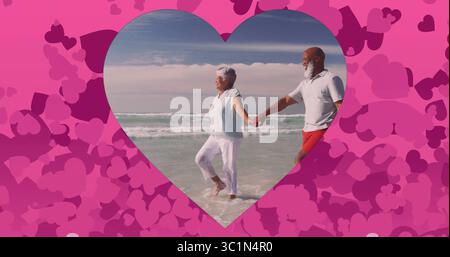 Walking senior couple holding hands in gentle waves at ocean shore, with pink heart frame overlay Stock Photo