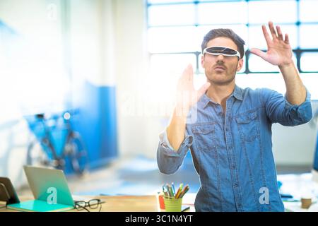Adult man standing behind wooden desk at modern studio wearing white VR glasses and using laptop Stock Photo