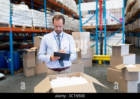 Male supervisor in office attire in warehouse holding clipboard inspecting box with packing peanuts Stock Photo