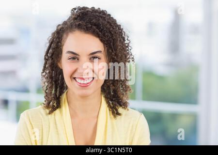 Woman smiling and standing in bright office lobby, showing blurred city skyline and green trees Stock Photo