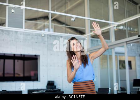 Woman gesturing toward virtual interface on glass partition in open-plan office with wall monitor Stock Photo