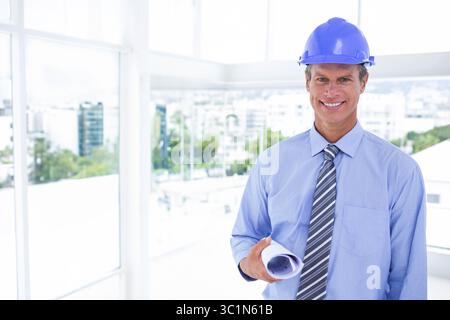 Middle-aged man wearing blue hard hat holding rolled blueprints standing inside office, copy space Stock Photo