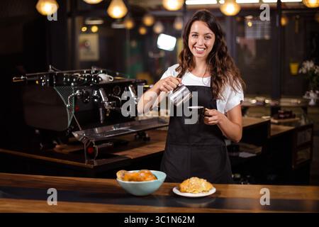 Inside cafe, steamed milk pouring from metal pitcher into coffee cup on plate with croissant Stock Photo