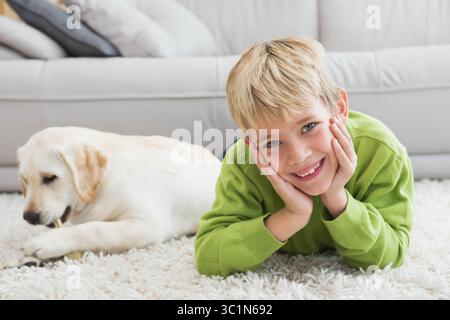 Blond boy lying on white shag rug at home smiling while puppy chewing bone near sofa Stock Photo