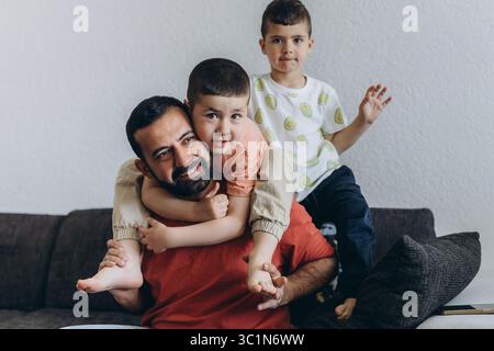 Happy middle eastern father having fun with his three sons at home. Dad cuddling toddler while other kids play and smile. Loving family moment on cozy Stock Photo