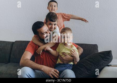 Happy middle eastern father having fun with his three sons at home. Dad cuddling toddler while other kids play and smile. Loving family moment on cozy Stock Photo