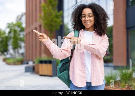 Smiling African American woman pointing to the side, wearing glasses, and carrying a backpack, in an urban setting. Stock Photo
