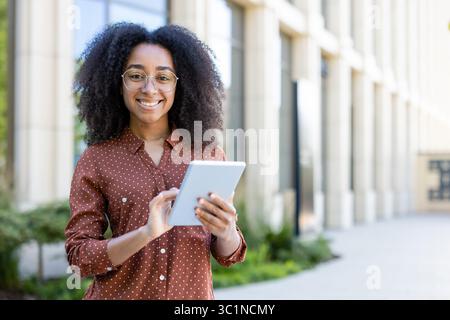 A young woman with glasses holds a tablet while smiling, with a blurred building in the background. Stock Photo