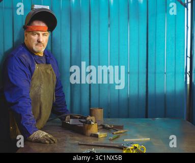 Welder wearing leather apron and helmet operating gas torch near cylinders in turquoise workshop Stock Photo
