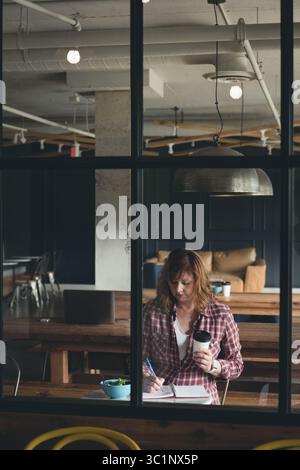 Red-haired woman taking notes and holding takeaway coffee cup beside salad bowl at workspace Stock Photo